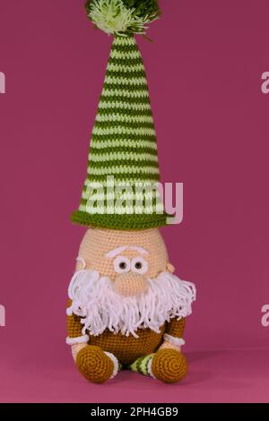 Amigurumi una nana con cappello a strisce verdi e barba bianca su sfondo rosa. Soffice giocattolo fai da te in cotone naturale e lana. Vecchio accovacciato, handma Foto Stock