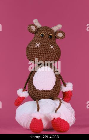 Amigurumi un toro marrone indossa guanti e ciabatte rossi e si siede su una palla bianca di filo su sfondo rosa. Soffice giocattolo fai da te in cotone naturale e w Foto Stock