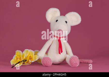 Amigurumi un mouse bianco con grandi orecchie e naso lungo siede su sfondo rosa accanto a fiori di orchidea gialla. Un morbido giocattolo fai da te in cotone naturale e. Foto Stock