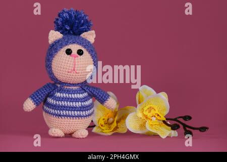 Bambola di gattino Amigurumi su sfondo rosa accanto a fiori di orchidea gialla. Un morbido giocattolo fai da te in cotone. Un gatto rosa che indossa abiti viola striati e. Foto Stock