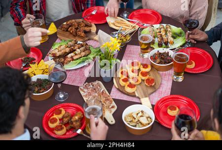 Un tavolo ben allestito che presenta una gamma di deliziose carni alla griglia, mini pizze, patatine croccanti bevande alcoliche. Gruppo multirazziale di amici hav Foto Stock