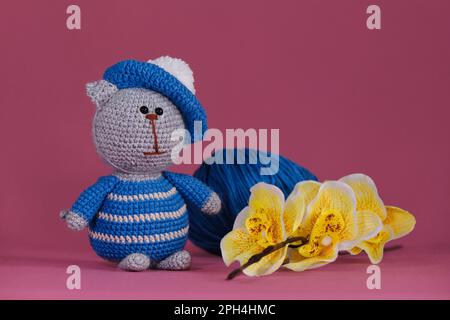 Bambola di gattino Amigurumi su sfondo rosa accanto a fiori di orchidea gialla. Un morbido giocattolo fai da te in cotone. Un gatto grigio che indossa abiti a strisce blu e. Foto Stock