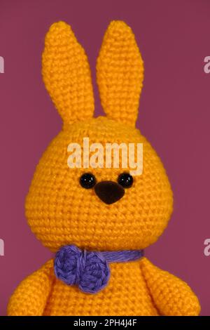 Simbolo della Pasqua cattolica. Coniglietto pasquale arancione con cravatta viola fianchettata, arte fatta a mano Bambola di coniglio Amigurumi su sfondo rosa. Giocattolo morbido fai da te Foto Stock