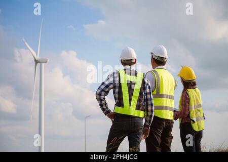Vista posteriore di tre ingegneri che discutono e controllano le turbine in una centrale di turbine eoliche. Tecnologia e sostenibilità delle energie rinnovabili. Foto Stock