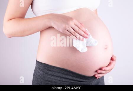 Pancia di donna incinta con calze bambino su sfondo grigio, vista dall'alto Foto Stock