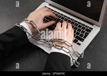 Dipendenza da Internet. Uomo che digita sul computer portatile con le mani incatenate al tavolo nero, primo piano Foto Stock