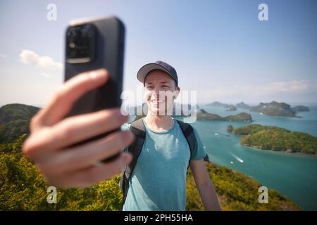 Uomo sorridente con zaino che porta un ritratto selfie in collina contro un gruppo di isole tropicali in mare. Ang Thong National Marine Park vicino a Koh Samui nel Th Foto Stock
