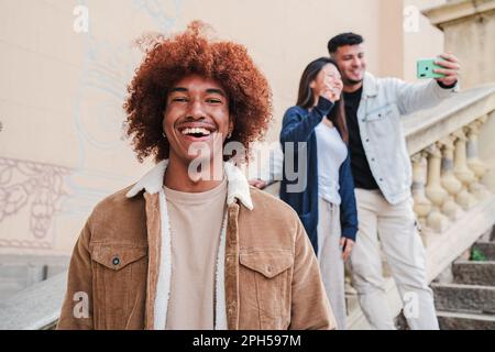In primo piano un bel giovane afro-americano sorridente e guardando la macchina fotografica con capelli afro, sullo sfondo due amici scattano un ritratto selfie. vista frontale di un ragazzo toothy maschio ridere. Foto di alta qualità Foto Stock