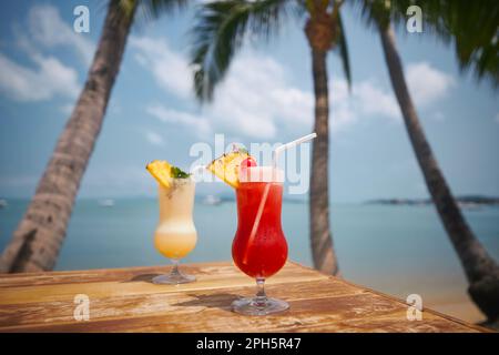 Due cocktail sotto le palme sulla splendida spiaggia. Drink Singapore Sling e Pina Colada su un tavolo di legno. Foto Stock