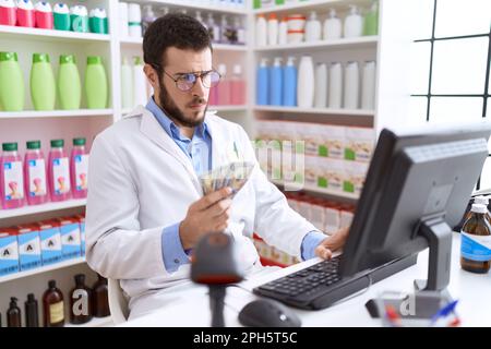 Farmacista giovane uomo ispanico che tiene dollari utilizzando il computer in farmacia Foto Stock