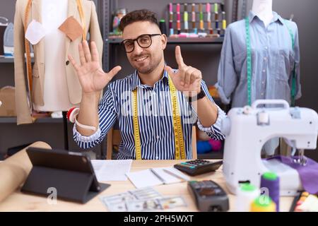 Uomo ispanico con Beard dresssaker designer che lavora presso atelier mostrando e indicando con le dita numero sette, mentre sorridendo fiducioso e felice. Foto Stock