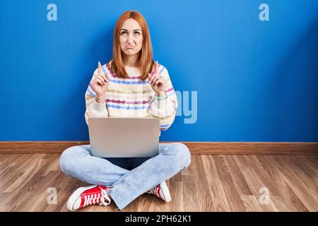 Giovane donna che usa il notebook a casa seduto sul pavimento, che indica un aspetto triste e sconvolto, indicando la direzione con le dita, infelice e depresso. Foto Stock