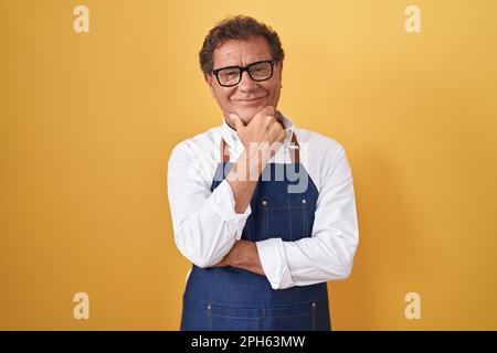 Uomo ispanico di mezza età che indossa grembiule cuoco professionale guardando sicuro la fotocamera sorridendo con braccia incrociate e la mano sollevata sul mento. pensare po Foto Stock