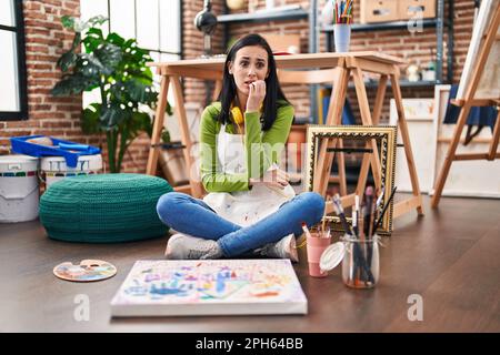 Donna ispanica seduta in studio di arte pittura su tela cercando stressato e nervoso con le mani su bocca mordente unghie. problema di ansia. Foto Stock