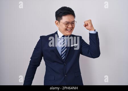 Giovane uomo asiatico che indossa il vestito di affari e danza cravatta felice e allegro, sorridente in movimento casuale e fiducioso ascoltare la musica Foto Stock