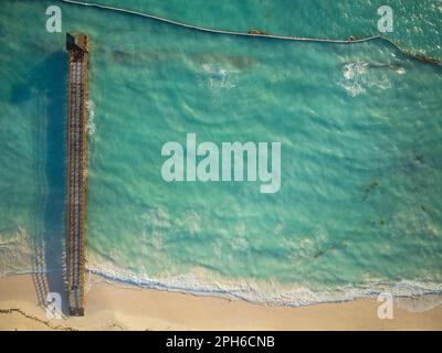 Vista dall'alto sul mare e su un molo in legno. Una leggera onda di mare corre sulla spiaggia sabbiosa. Minimalismo. Ecologia, clima, tempo, protezione ambientale. Foto Stock
