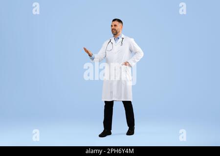 Medico di mezza età che mostra spazio copia con la mano, indossare uniforme, camminare su sfondo blu studio, a tutta lunghezza Foto Stock