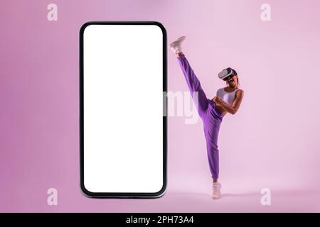 Adatta la ragazza afro-americana in googles vr esplorare il gioco di realtà virtuale vicino al grande smartphone con schermo vuoto, mockup Foto Stock