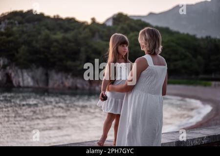 Primo piano ritratto di madre incinta con bambina che cammina mano in mano in abiti bianchi al tramonto sul mare. Assistenza ai bambini. Maternità sana. Foto Stock