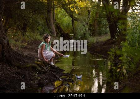 Ragazza adolescente con capelli verdi in vintage vestito, si trova sulla riva del fiume, lancia carta bianca origami barca in acqua nella foresta. Traslazione concep Foto Stock