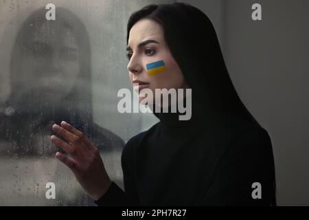 Rovesciato giovane donna con la foto della bandiera Ucraina sulla guancia vicino alla finestra a casa. Fermare la guerra in Ucraina Foto Stock