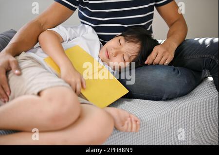Rilassato e felice giovane figlio asiatico ascoltò una fiaba e si addormentò in grembo del padre sul divano del soggiorno. Foto Stock