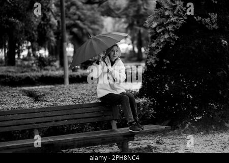 Una donna con un ombrello siede su una panchina nel parco. Foto in bianco e nero. Foto Stock