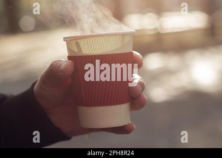 una mano dell'uomo tiene una tazza di carta con tè caldo sulla strada nel parco nel pomeriggio al sole Foto Stock