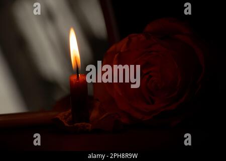 una fiamma di candela e accanto ad una rosa nel primo piano scuro Foto Stock