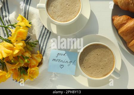 Caffè, croissant, fiori e scheda con frase BUONGIORNO! su tavolo di legno bianco, piatto Foto Stock