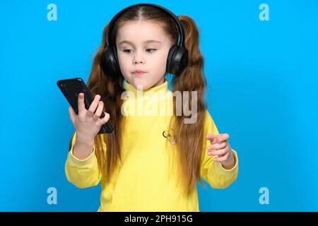 Ivano-Frankivsk, Ucraina 28 gennaio 2022: Bella bambina utilizza cuffie wireless jbl e xiomi telefono cellulare, ritratto di un bambino su un blu b Foto Stock