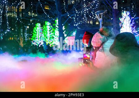 Persone che scattano foto di luci colorate installate presso le fontane di Jubille Park durante Canary Wharf Winter Lights 2023, Londra, Regno Unito Foto Stock