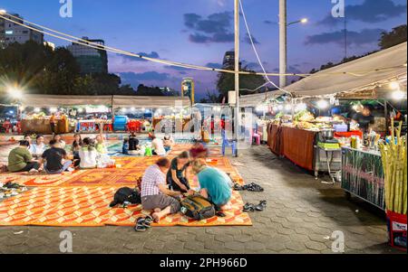 Phnom Penh, Cambogia-Dicembre 23rd 2022: Come le cascate del crepuscolo, Khmer e viaggiatori occidentali sedersi in piccoli gruppi su stuoie, in una grande area, circondato da bancarelle alimentari Foto Stock