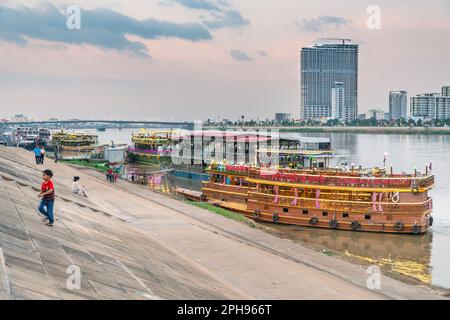 Phnom Penh, Cambogia-Dicembre 23rd 2022: Al crepuscolo, gli abitanti del posto camminano lungo le rive del fiume Tonle SAP, passato queste imbarcazioni vivacemente illuminate, utilizzato come locali notturni e. Foto Stock