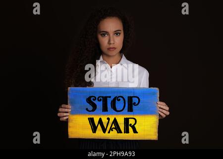 Donna afro-americana infelice con segno con frase Stop War su sfondo nero Foto Stock