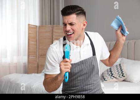Uomo con spazzola e straccio che canta mentre pulisce in camera da letto Foto Stock