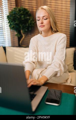 Immagine verticale di una giovane ragazza bionda attraente che utilizza un computer portatile in un moderno bar, guardando lo schermo con un look concentrato Foto Stock