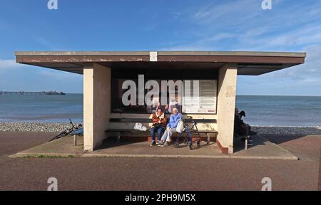Due Signore che godono di un soleggiato rifugio dal vento, Llandudno Promenade, Conwy, Galles del Nord, Regno Unito, LL30 2LP Foto Stock