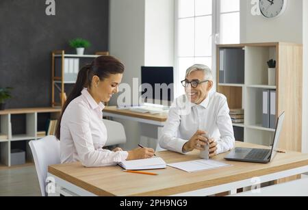Un senior manager sorridente che consulti un giovane dipendente seduto alla scrivania dell'ufficio Foto Stock
