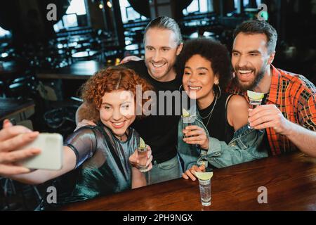 Allegri amici interrazziali con tequila scatti selfie su smartphone in bar, immagine stock Foto Stock