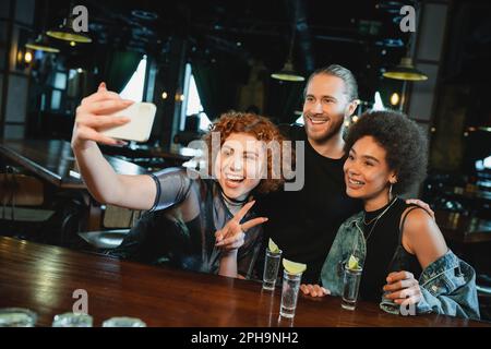 Allegri amici multietnici che prendono selfie su smartphone vicino tequila in bar, immagine stock Foto Stock