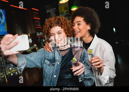 Fidanzata amici multietnici prendere selfie e tequila tenere in bar, immagine stock Foto Stock