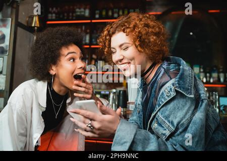 Donna eccitata redhead utilizzando smartphone vicino afroamericano amico in bar, immagine stock Foto Stock