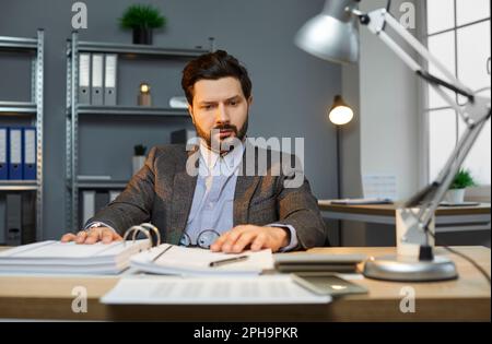Giovane uomo d'affari attraente con barba e baffi in tuta che lavora alla scrivania sul suo posto di lavoro Foto Stock