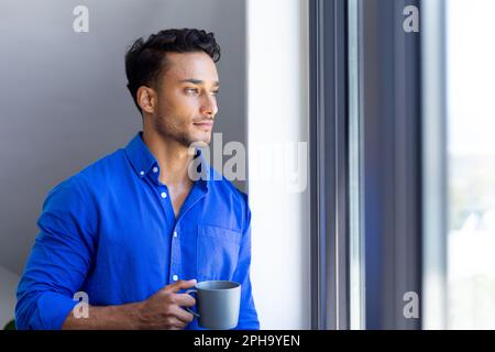 Felice uomo biraciale tenendo la tazza e guardando fuori dalla finestra Foto Stock