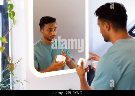 Uomo biraciale che guarda se stesso nello specchio del bagno e applica crema viso Foto Stock