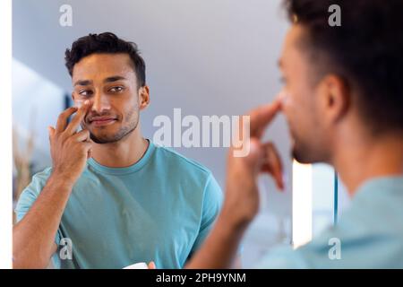 Uomo biraciale sorridente che guarda se stesso nello specchio del bagno e applica crema viso Foto Stock