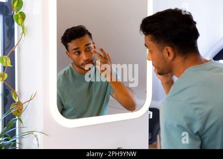 Uomo biraciale che si guarda nello specchio del bagno e si applica sotto la crema per gli occhi Foto Stock