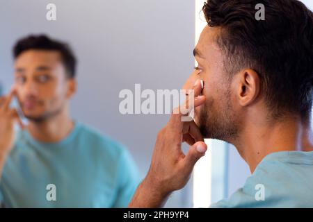 Uomo biraciale che guarda se stesso nello specchio del bagno e applica crema viso Foto Stock