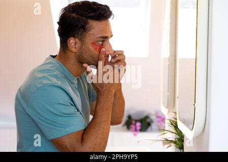 Uomo biraciale che guarda nello specchio e che applica sotto la maschera oculare in bagno Foto Stock